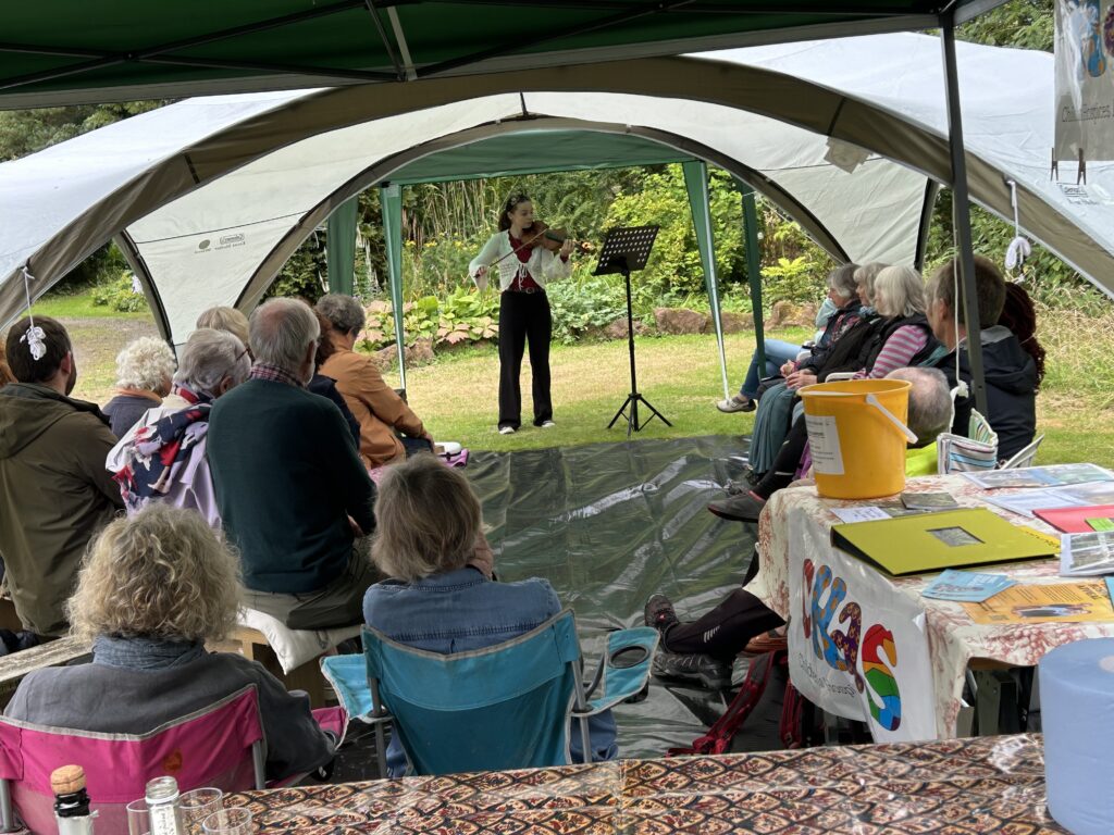 Ola Stanton plays the violin in the gazebo framed by plants at Pond Cottage. The audience sits in appreciation of a beautiful performance